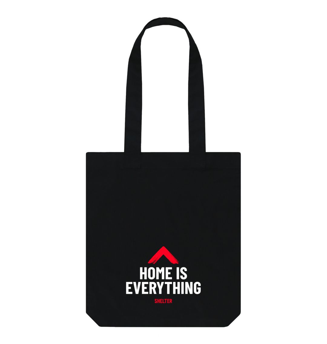 A black tote bag with the slogan 'Home is Everything' in white font and the Shelter roof logo in red above the text