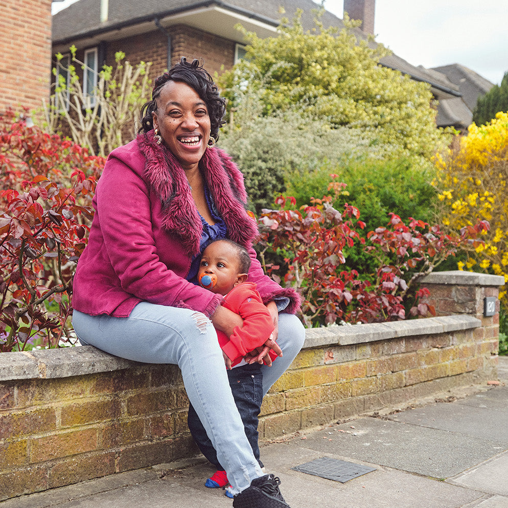 A woman sitting on a brick wall outdoors, holding a baby. She is wearing a magenta zip-up hoodie, a dark pink scarf, and light blue jeans.