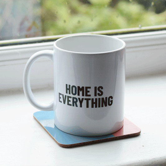A white ceramic mug on a window sill. The mug reads 'Home is Everything' on one side and has the Shelter logo on the reverse.