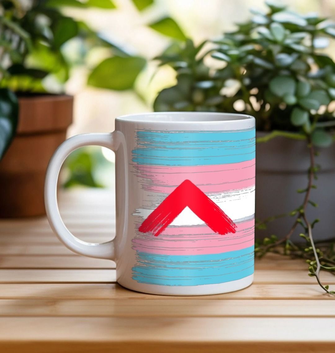 A white ceramic mug with the trans flag colours and the Shelter logo on the reverse