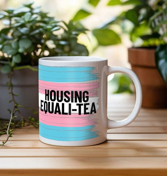 A white ceramic mug with the words 'Housing Equali-tea' over the trans flag colours. 