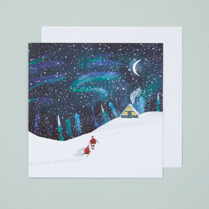 A square Christmas card with an  atmospheric, Nordic inspired illustration showing Father Christmas pulling a sled full of presents ready to deliver to a cosy cottage on a snowy Christmas Eve.