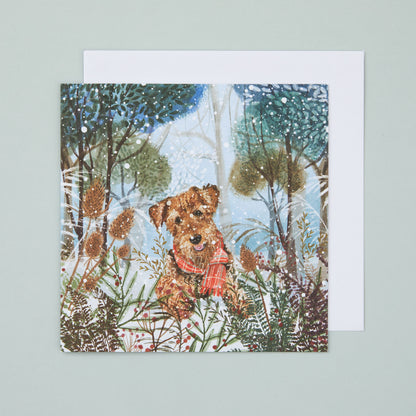 A square Christmas card with an illustration depicting an airedale terrier in a snowy woodland scene.