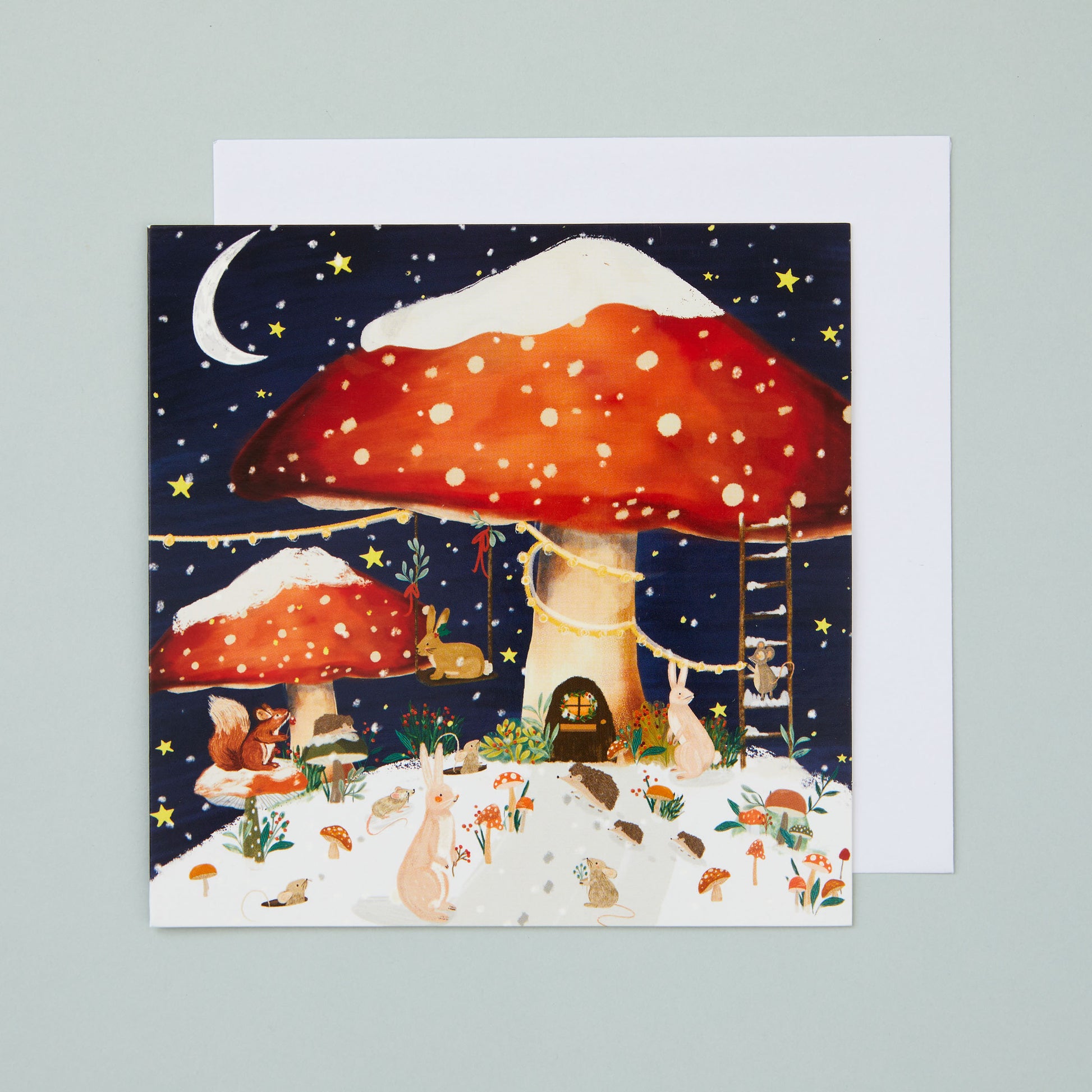 A square Christmas card with an illustrated whimsical winter scene with fairytale-esque toadstool houses and a collection of cute woodland animals, all under a starry night sky.