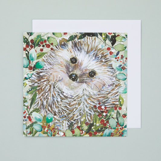 A square Christmas card with an illustration depicting a hedgehog surrounded by green leaves and red berries.