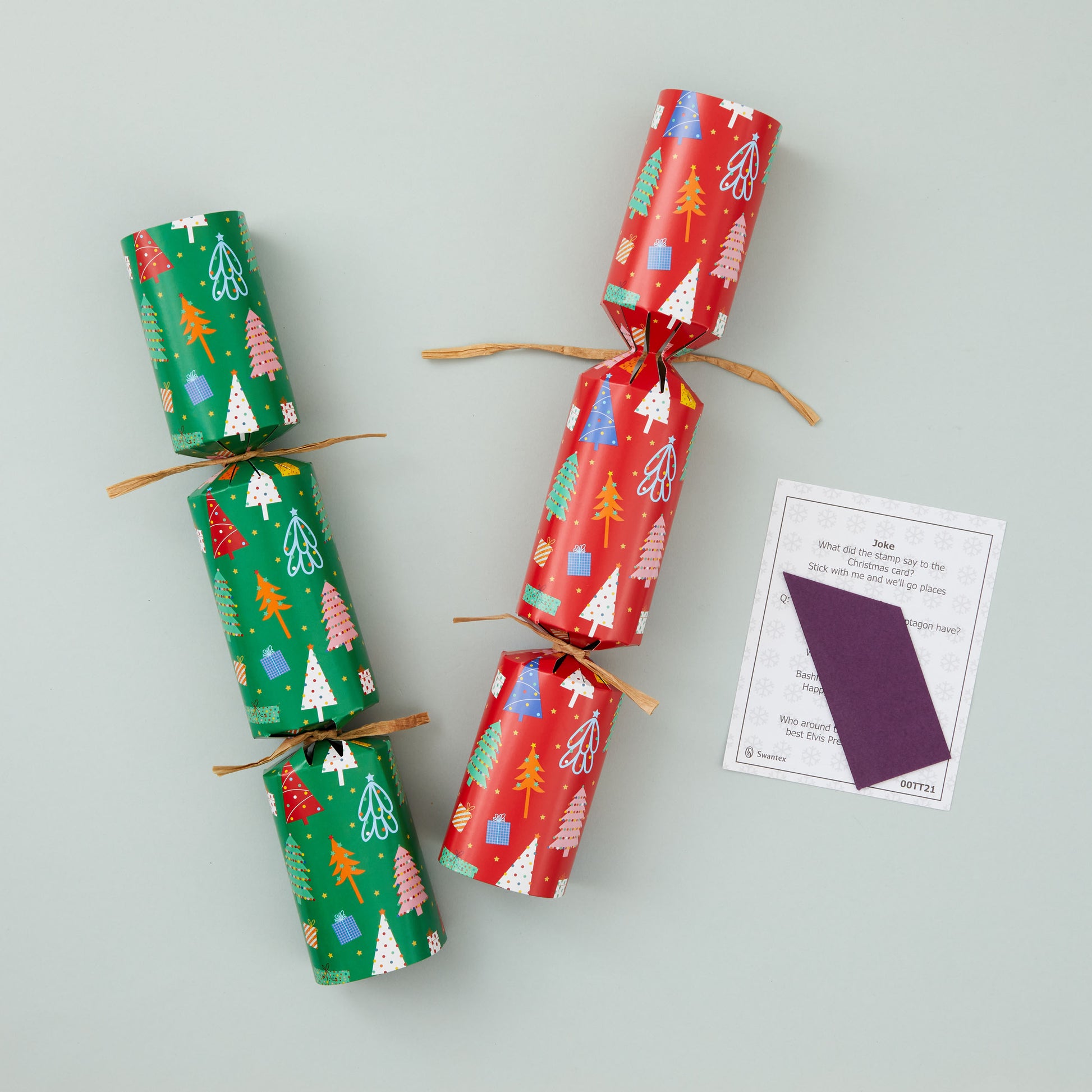 Two Christmas crackers with red and green Christmas tree design with folded paper hat and joke.