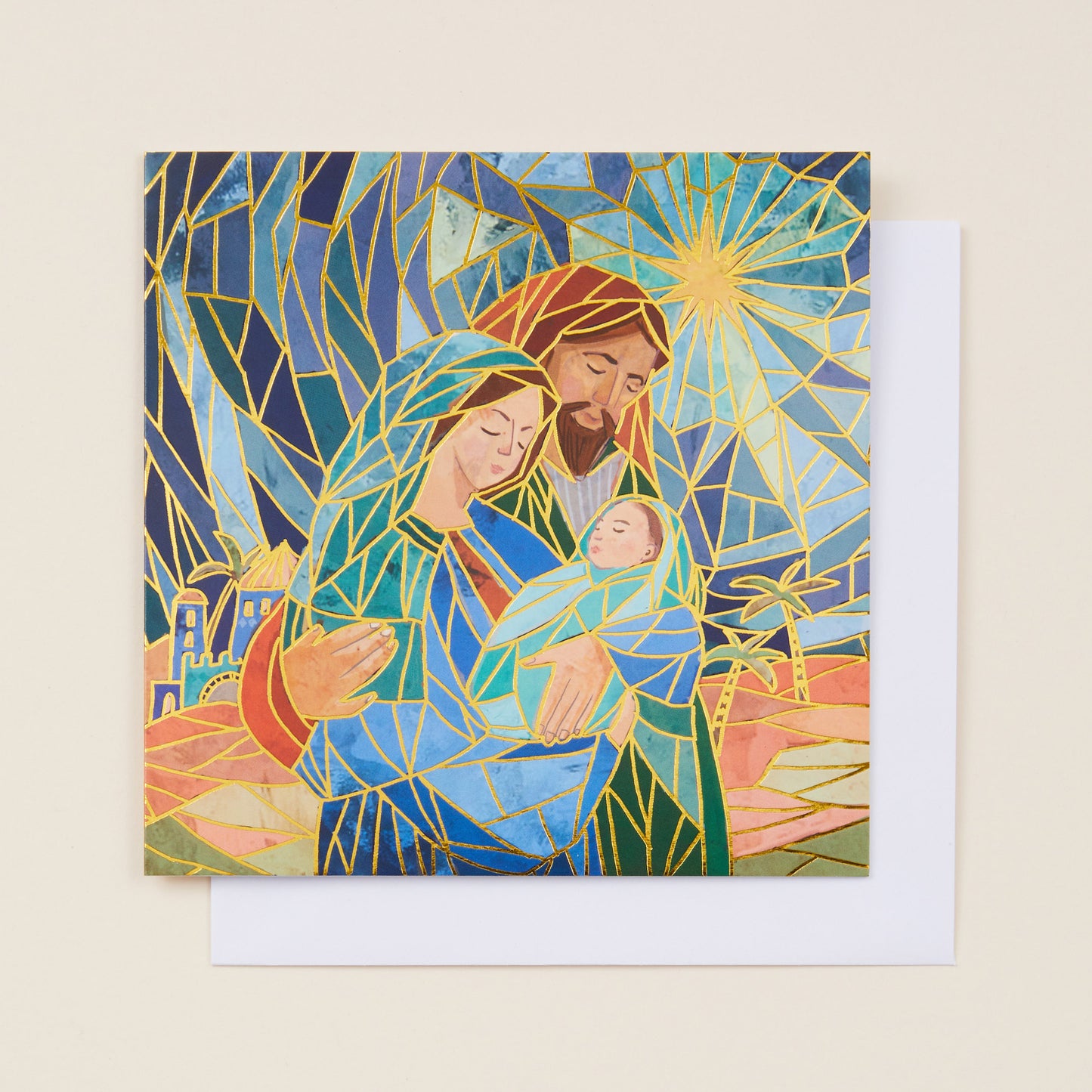 A square Christmas card with a stylized image of Mary, Joseph and Jesus in Bethlehem on the front, the card sitting on a white envelope.