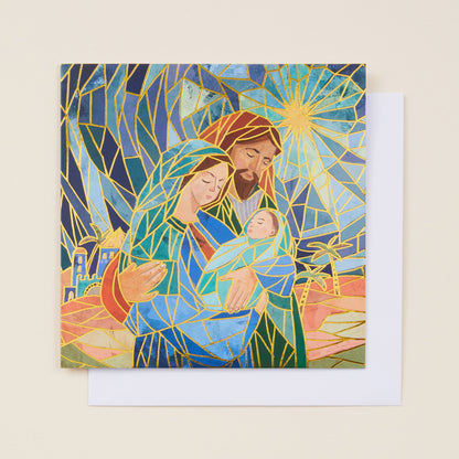 A square Christmas card with a stylized image of Mary, Joseph and Jesus in Bethlehem on the front, the card sitting on a white envelope.