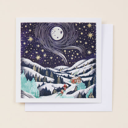 A square Christmas card with a snowy mountain scene on the front featuring Santa, the card sitting on a white envelope.