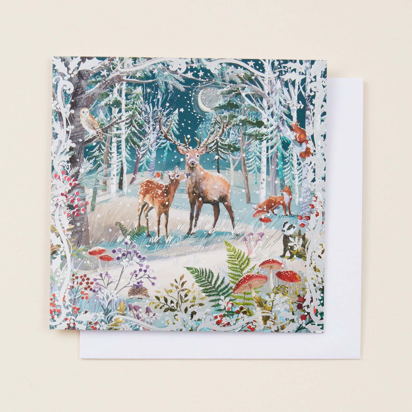 A square Christmas card with a snowy woodland scene on the front, sitting on a white envelope.
