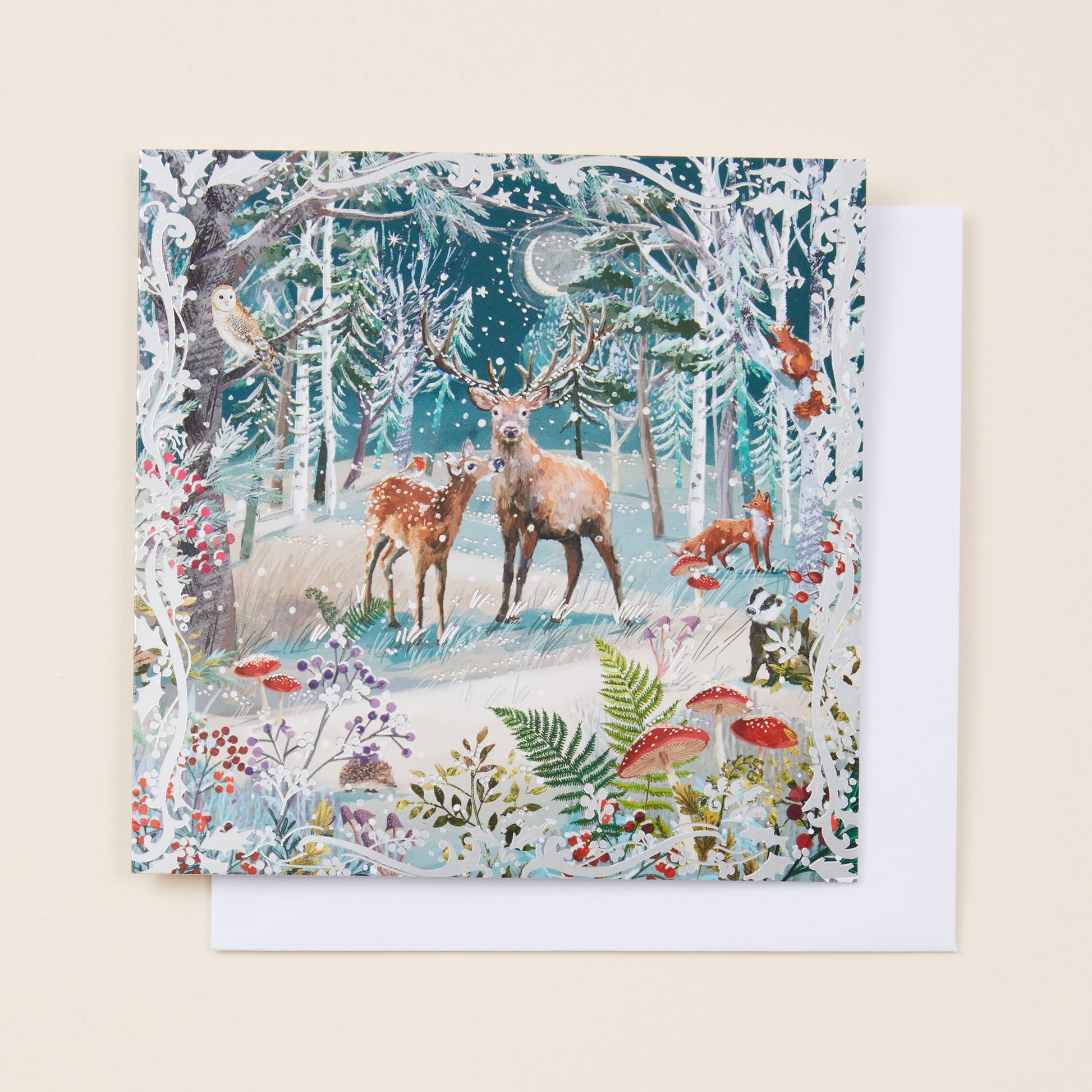 A square Christmas card with a snowy woodland scene on the front, sitting on a white envelope.