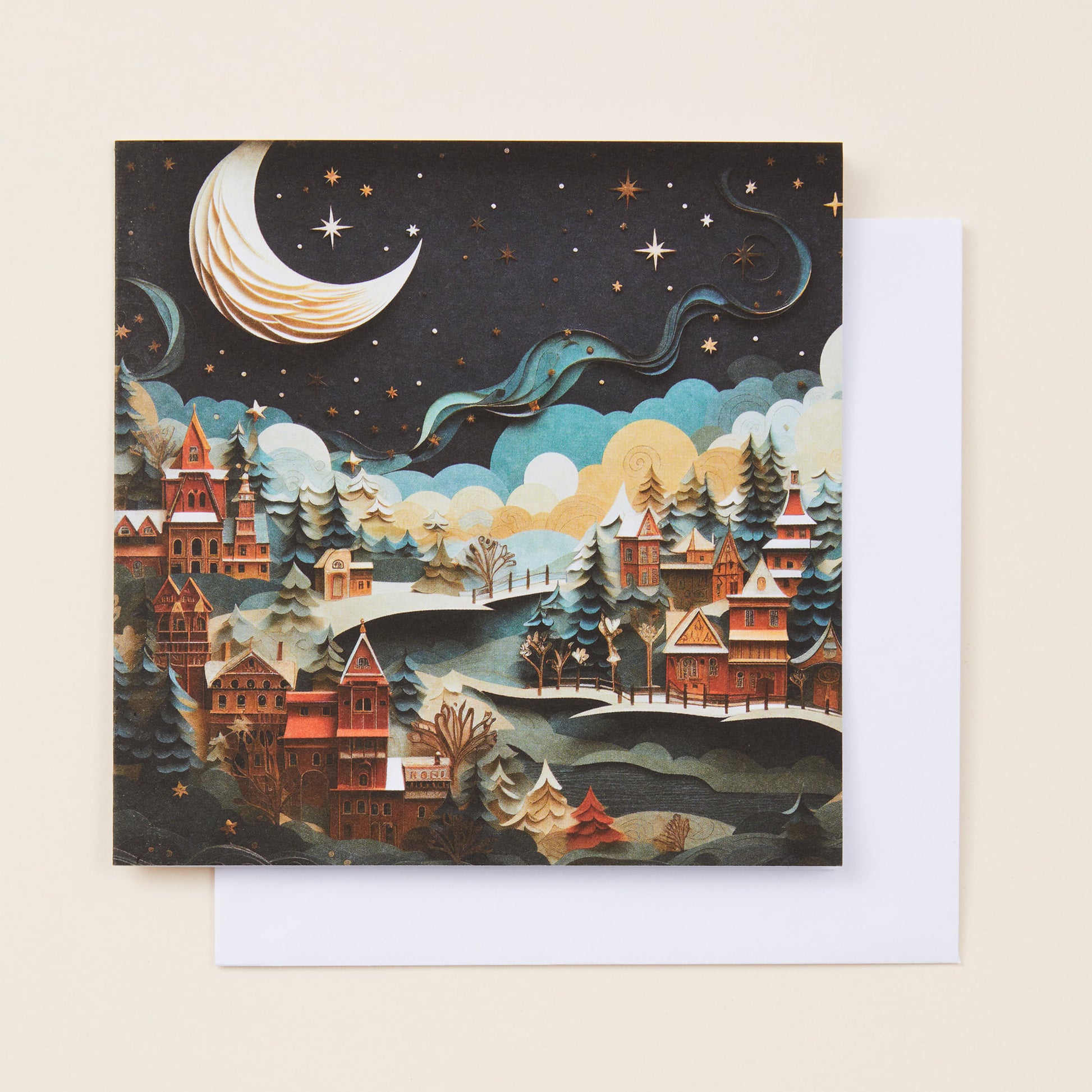 A square Christmas card with a paper-like stylised image on the front of a town under a crescent moon, sitting on a white envelope