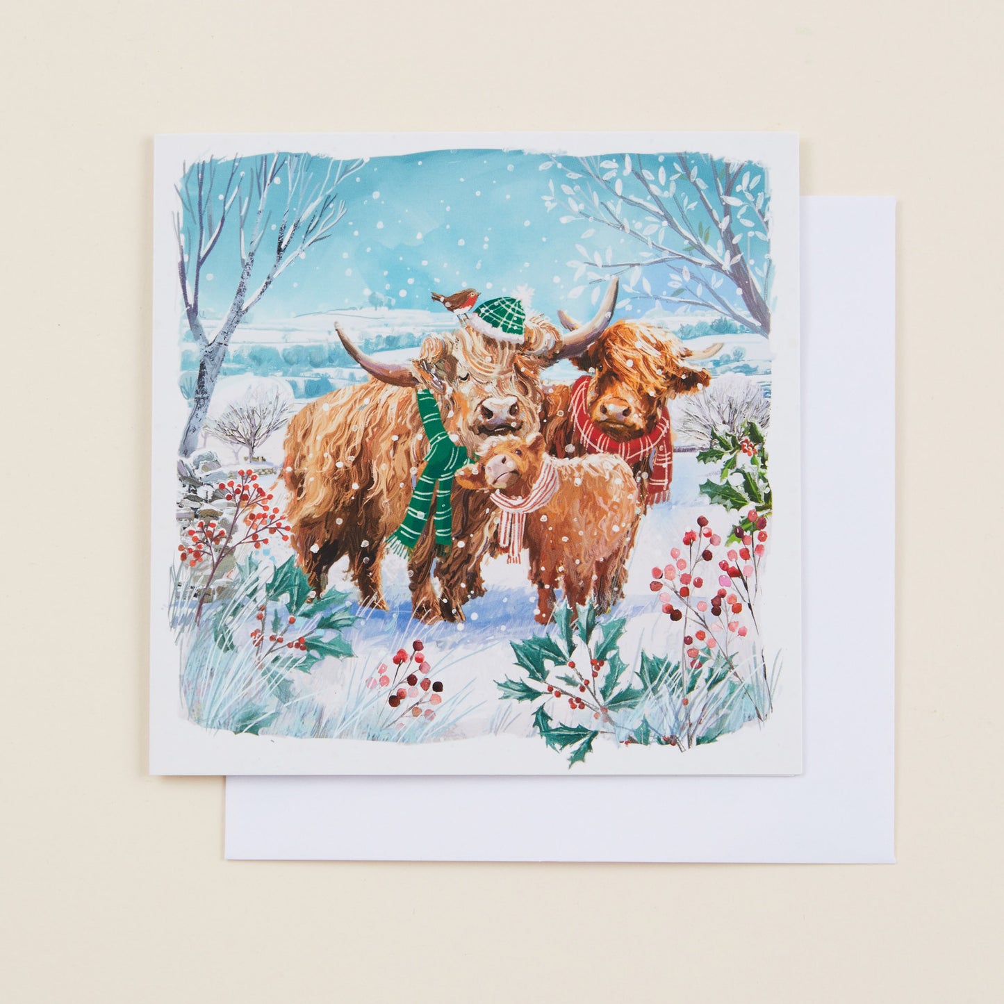 A square Christmas card with an illustration of highland cows wearing scarves and hats on the front, sitting on a white envelope.