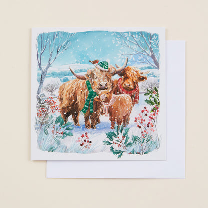 A square Christmas card with an illustration of highland cows wearing scarves and hats on the front, sitting on a white envelope.