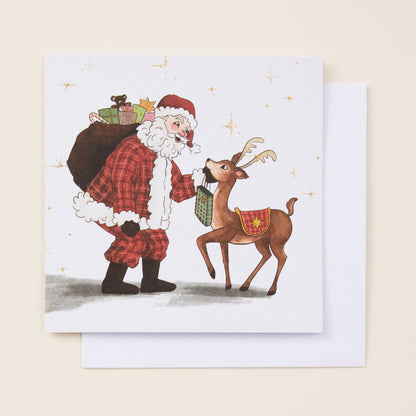 A square Christmas card with a cartoon illustration on the front featuring Santa and a reindeer. The card is sitting on a white envelope.