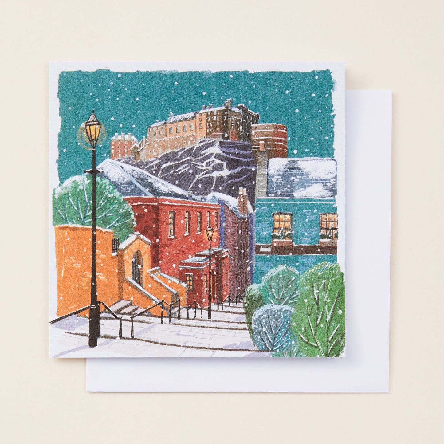 A square Christmas card showing a snowy town underneath Edinburgh Castle, sitting on a white envelope.