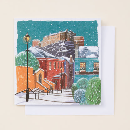 A square Christmas card showing a snowy town underneath Edinburgh Castle, sitting on a white envelope.