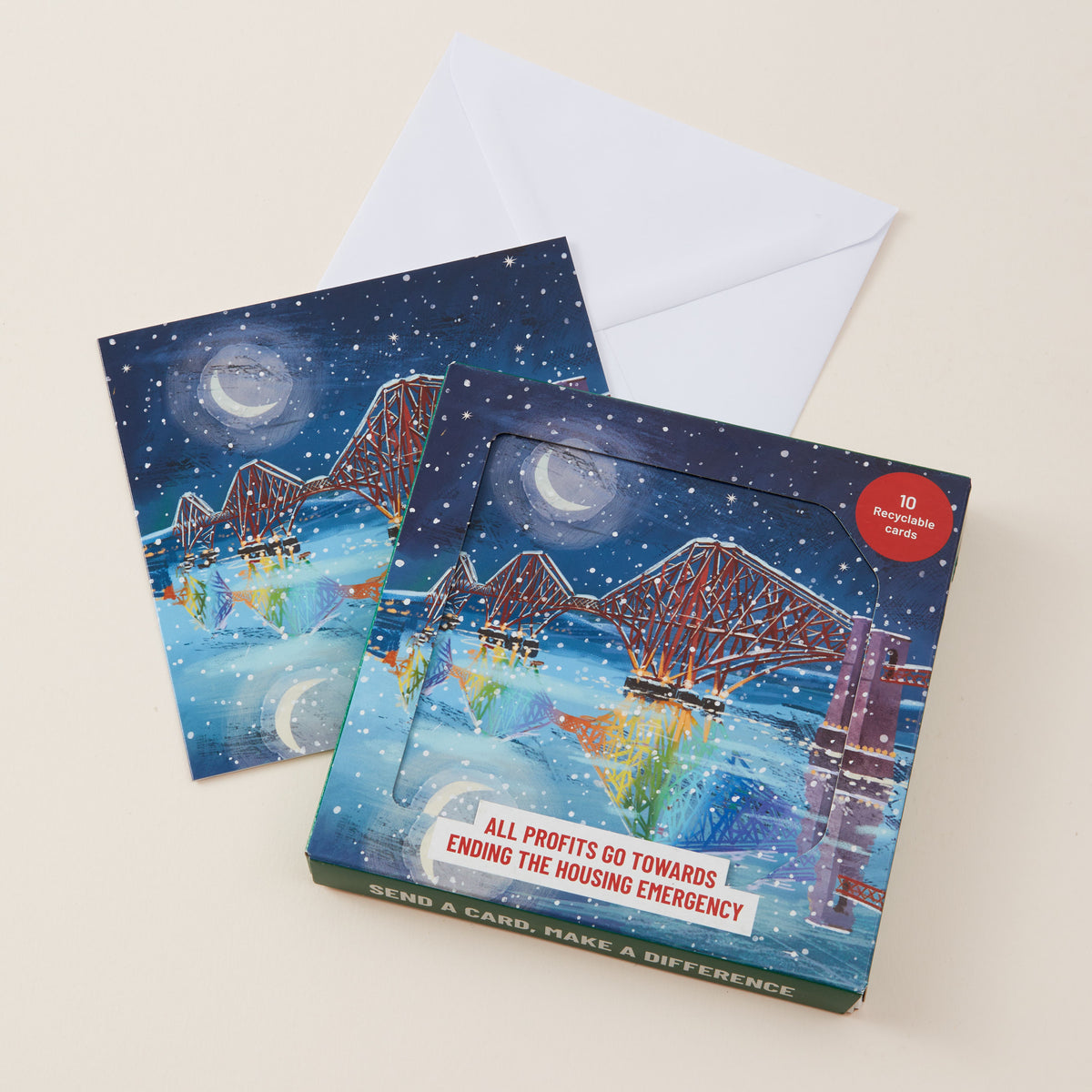 Two illustrated greeting cards with a white envelope behind them, photographed on a light beige background. The front card shows a night city scene with a depiction of the Forth Bridge, moon, and colorful reflections, plus a red circular badge reading '10 inspirational cards' and a slogan near the bottom.