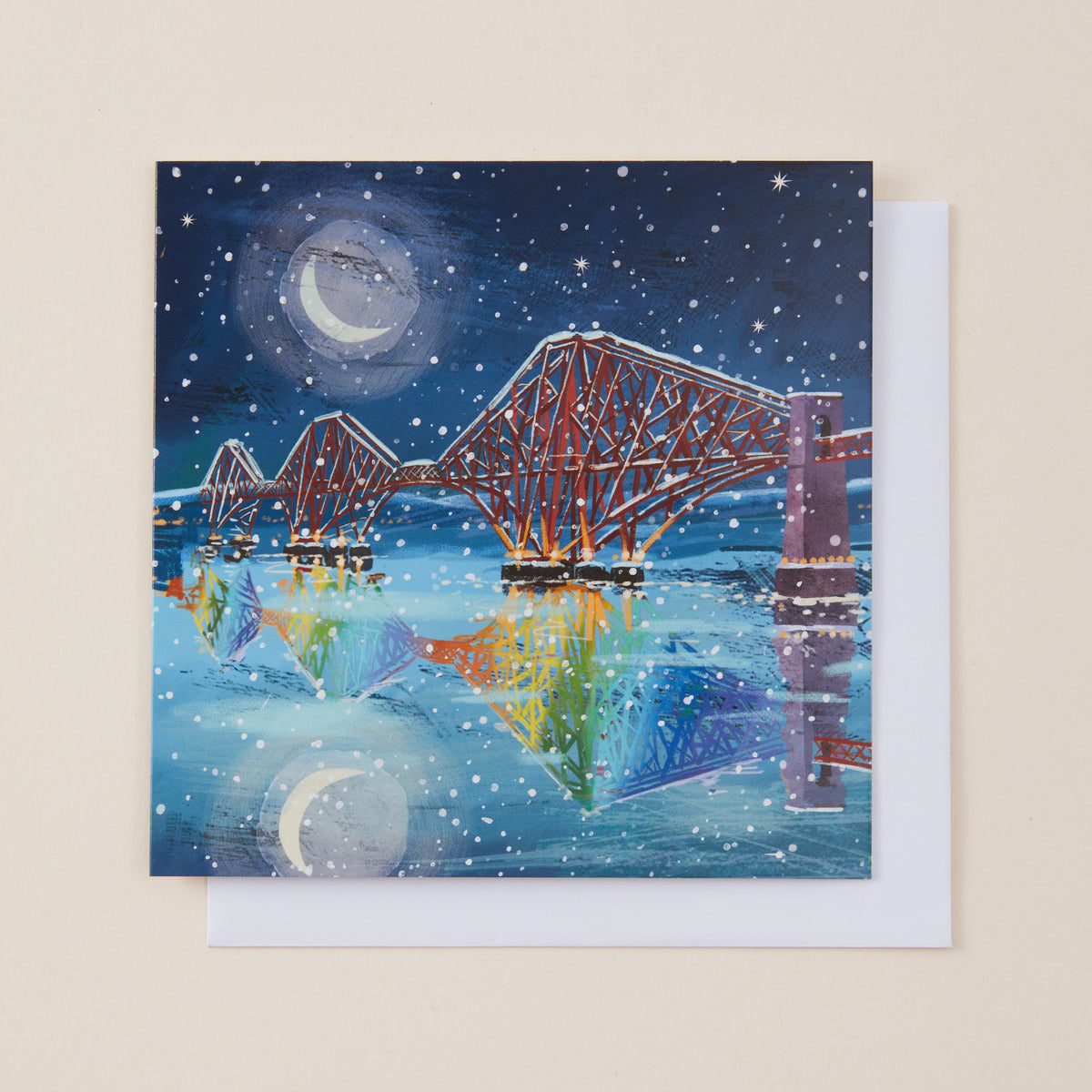 A square art print or card featuring a snowy night scene with a red-orange depiction of the Forth Bridge over reflective water under a crescent moon and stars, shown with a white envelope behind it on a light beige background.