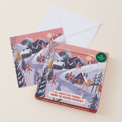 A boxed holiday card set with two illustrated winter mountain cards, a white envelope, and a red box. The front of the box shows a snowy village scene with cabins, trees, and a green circular badge that reads '10 Christmas cards.