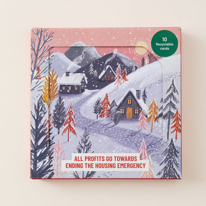 A square product box featuring a winter village illustration with snowy hills, cabins, trees, and mountains. The box has a pink border, a green circular badge reading '10 Recyclable cards,' and a white banner near the bottom that says 'ALL PROFITS GO TOWARDS ENDING THE HOUSING EMERGENCY.