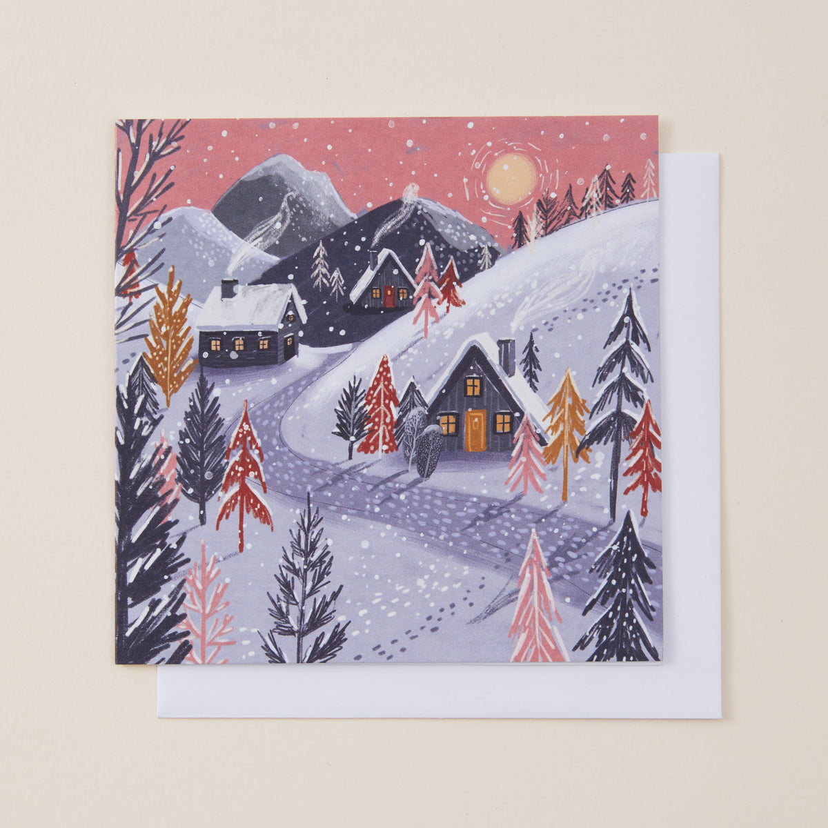 A square winter landscape Christmas card featuring snow-covered mountains, cabins, evergreen trees, and a pink sky with a moon, shown with a white envelope behind it on a light beige background.