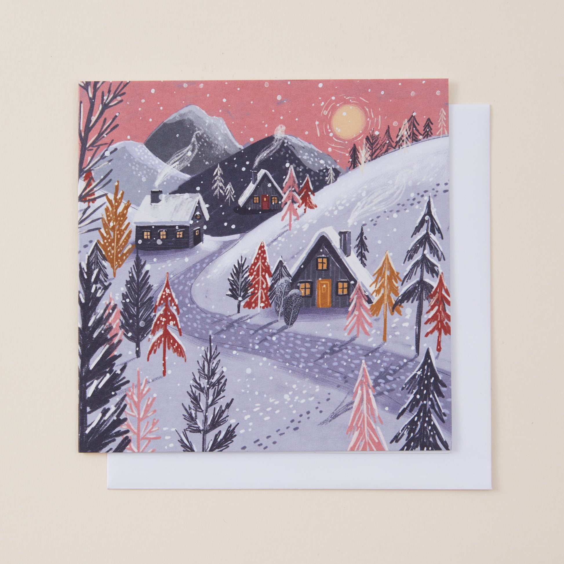 A square winter landscape Christmas card featuring snow-covered mountains, cabins, evergreen trees, and a pink sky with a moon, shown with a white envelope behind it on a light beige background.