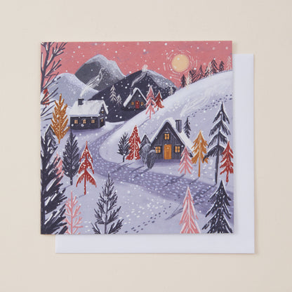 A square winter landscape Christmas card featuring snow-covered mountains, cabins, evergreen trees, and a pink sky with a moon, shown with a white envelope behind it on a light beige background.