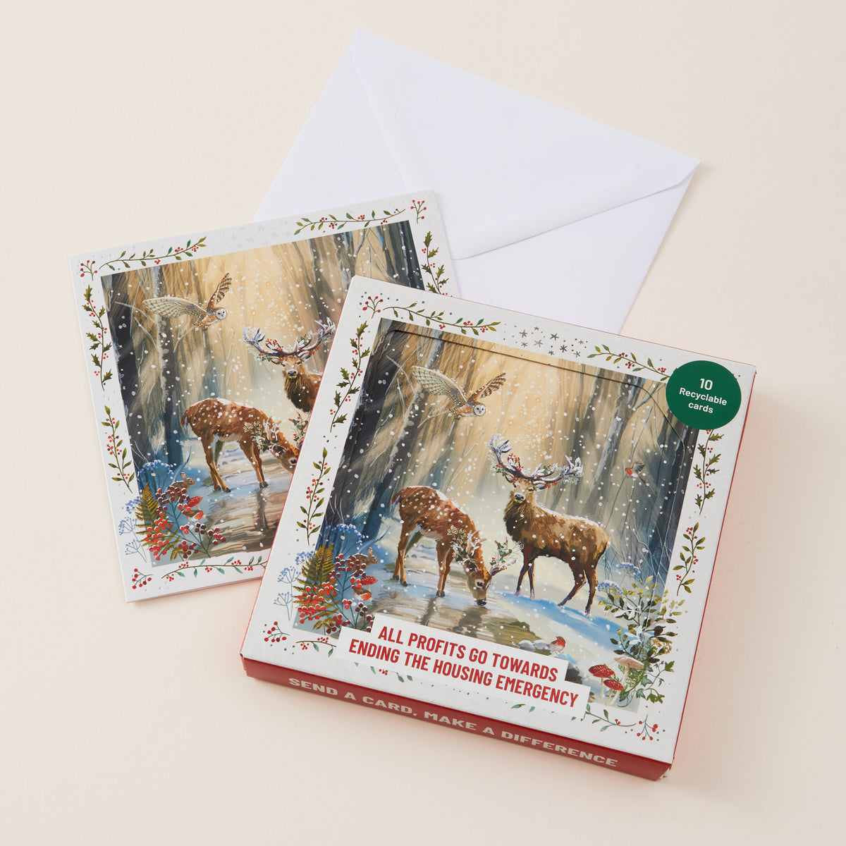 Two illustrated greeting cards with a white envelope, shown on a light beige background. The front card features a snowy forest scene with deer and birds.