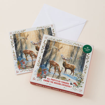 Two illustrated greeting cards with a white envelope, shown on a light beige background. The front card features a snowy forest scene with deer and birds.