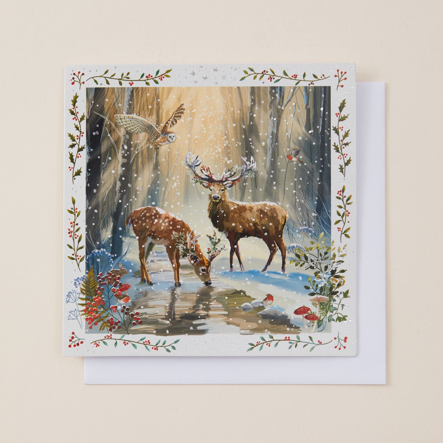 A square greeting card with a white envelope, featuring a winter woodland illustration of two deer, a bird, and a snowy forest scene, set against a warm beige background.