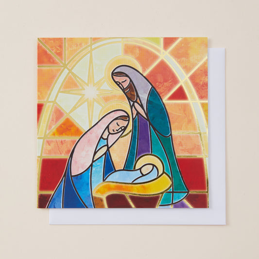 A square greeting card featuring a stained-glass style illustration of Mary, Joseph, and baby Jesus in warm orange, yellow, red, blue, and purple tones, shown with a white envelope on a light beige background.