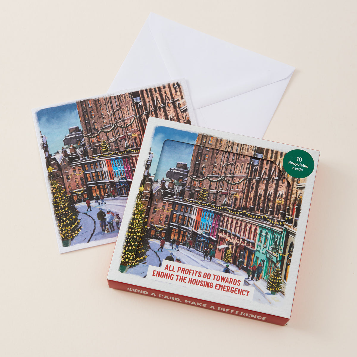 A boxed card set with two illustrated holiday city street cards and a white envelope on a light beige background. The front box shows a snowy urban street scene with buildings, Christmas trees, and red text reading 'SEND A CARD, MAKE A DIFFERENCE' and 'ALL PROFITS GO TOWARDS ENDING THE HOUSING EMERGENCY.