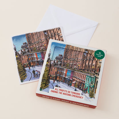 A boxed card set with two illustrated holiday city street cards and a white envelope on a light beige background. The front box shows a snowy urban street scene with buildings, Christmas trees, and red text reading 'SEND A CARD, MAKE A DIFFERENCE' and 'ALL PROFITS GO TOWARDS ENDING THE HOUSING EMERGENCY.