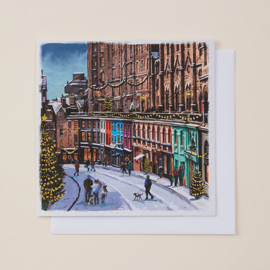 A square art print or greeting card featuring a colorful winter city street scene with snow, buildings, pedestrians, and decorated trees, shown with a white envelope on a light beige background.