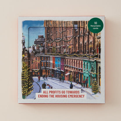 A square printed card or card set box featuring an illustrated snowy city street scene with colorful buildings and small figures. A green circular badge in the top right says '10 Recyclable cards,' and a white banner at the bottom reads 'ALL PROFITS GO TOWARDS ENDING THE HOUSING EMERGENCY.