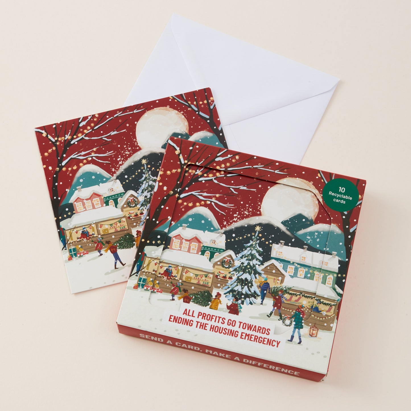 A Christmas-themed card box with a white envelope behind it, photographed on a light beige background. The box features a winter village illustration in red, white, blue, and green tones..