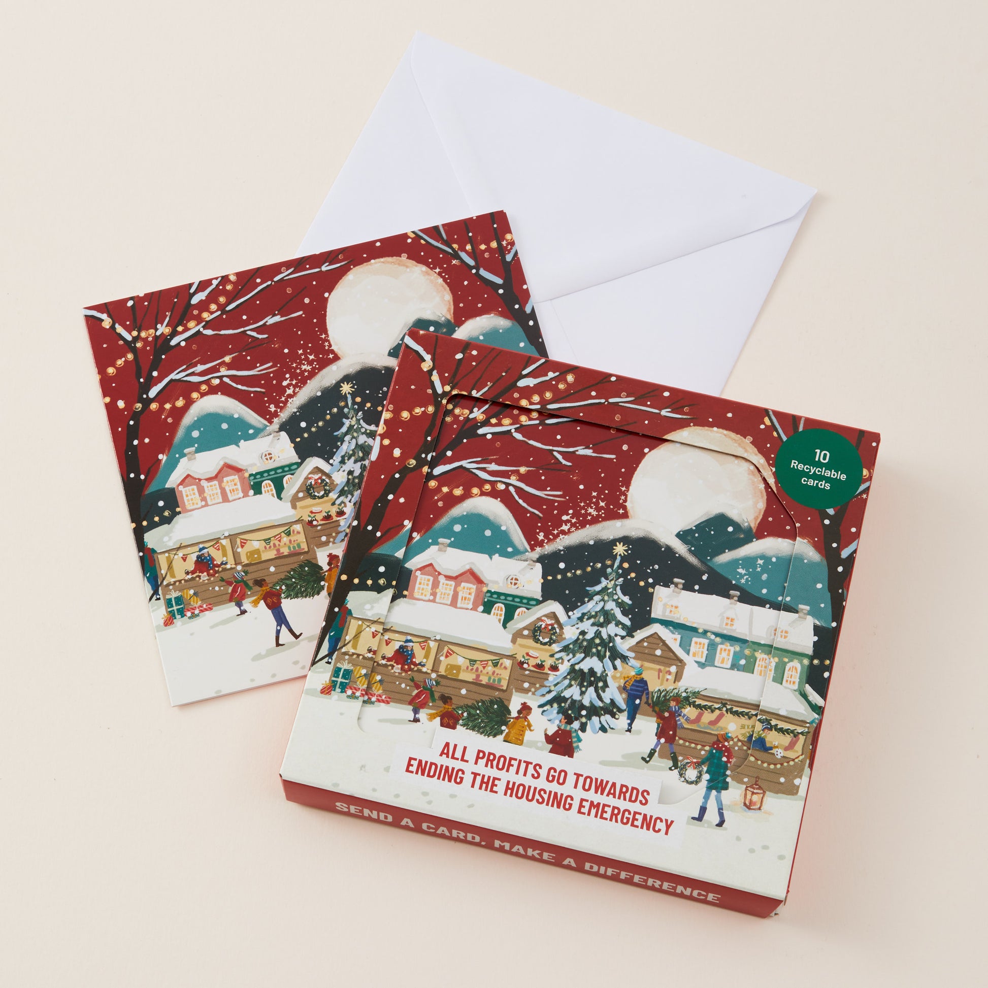 A Christmas-themed card box with a white envelope behind it, photographed on a light beige background. The box features a winter village illustration in red, white, blue, and green tones..