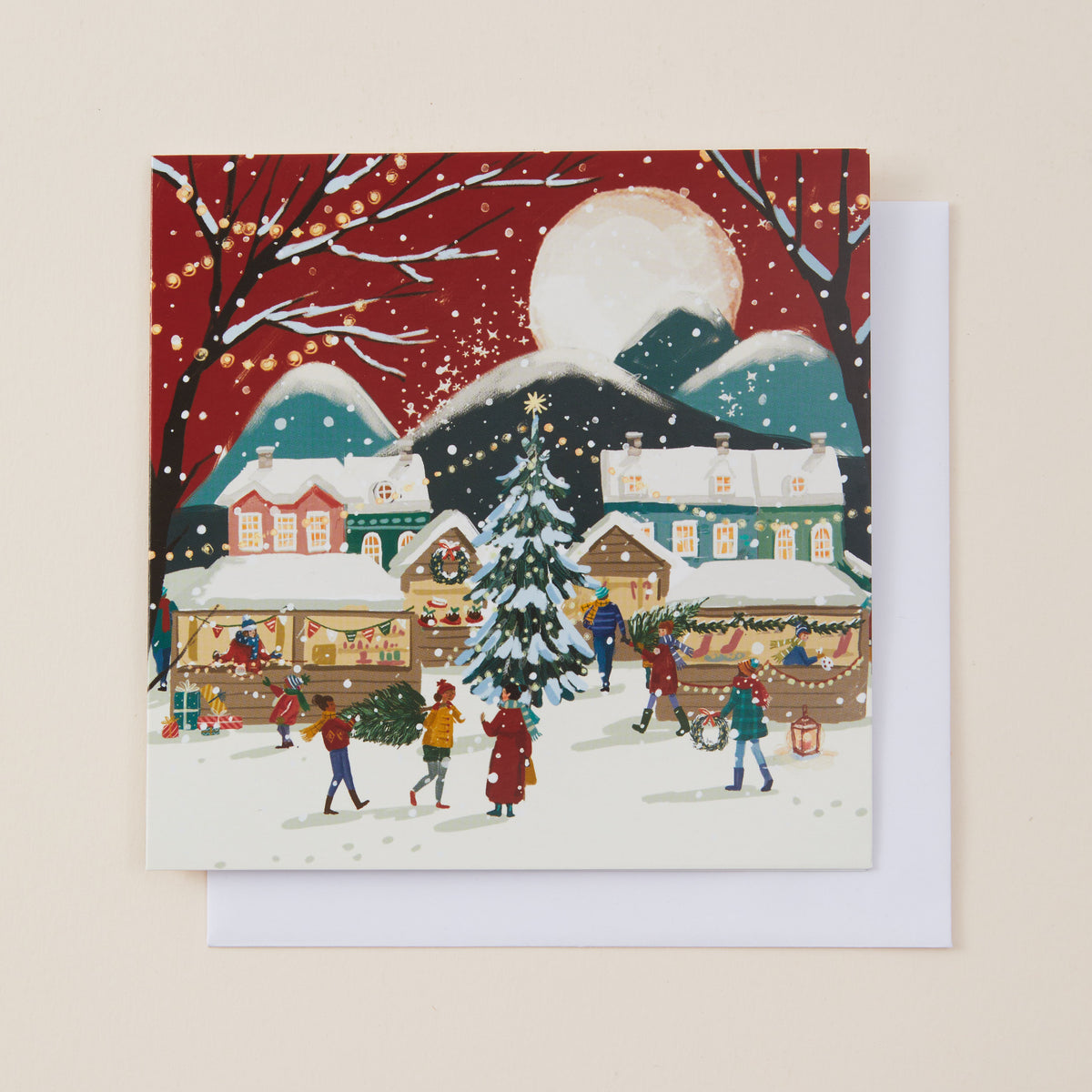A square Christmas greeting card featuring a festive village scene with snow-covered houses, a decorated Christmas tree, people, and a large moon, shown with a white envelope behind it on a light beige background.