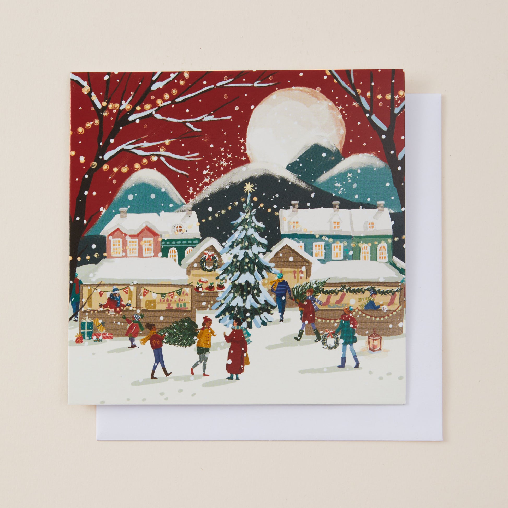 A square Christmas greeting card featuring a festive village scene with snow-covered houses, a decorated Christmas tree, people, and a large moon, shown with a white envelope behind it on a light beige background.