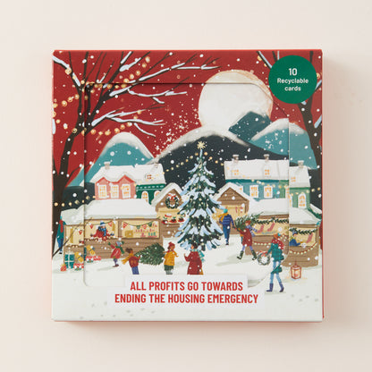 A square illustrated product box shown against a light beige background. The front artwork depicts a snowy village scene at night with houses, a Christmas tree, people, falling snow, and a red sky.