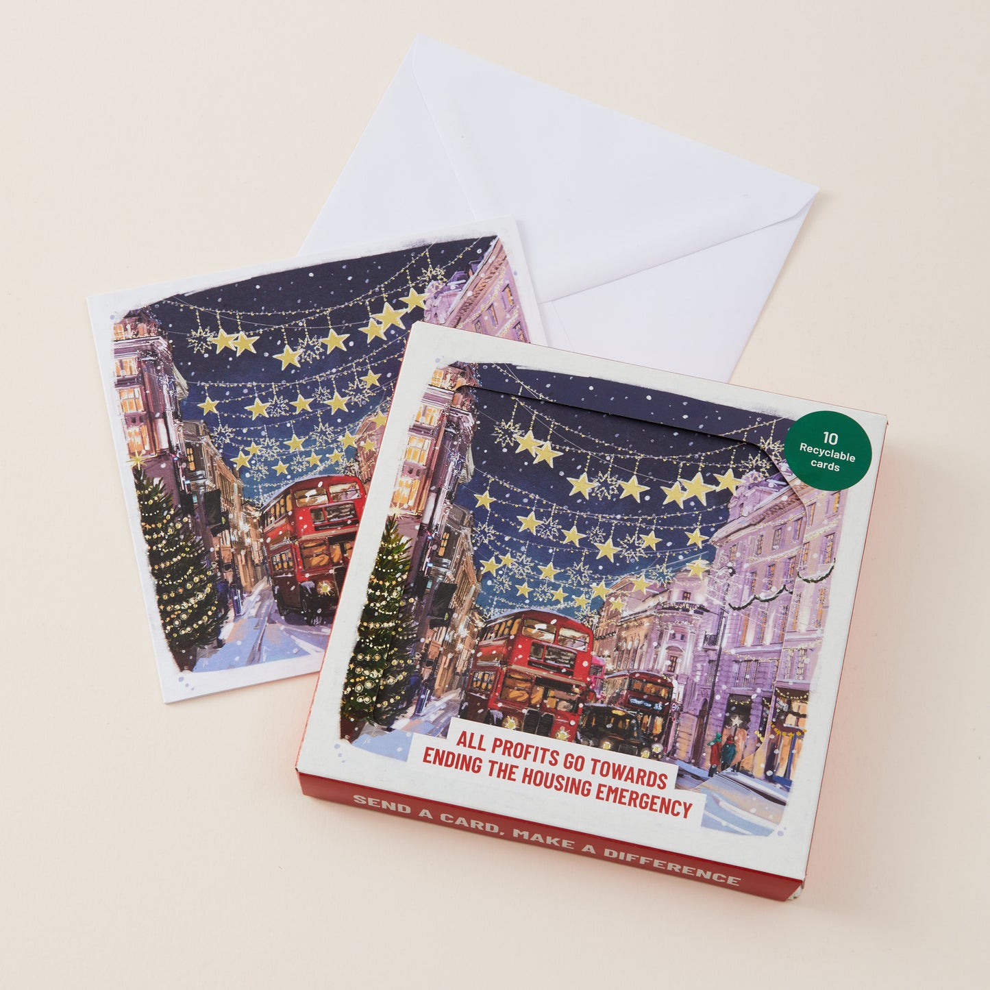 A boxed holiday card set with two illustrated cards and white envelopes on a light beige background. The front box shows a snowy city street scene with red buses, decorated trees, and starry night sky, plus text reading 'ALL PROFITS GO TOWARDS ENDING THE HOUSING EMERGENCY' and a green circular badge that says '10 illustrated cards.