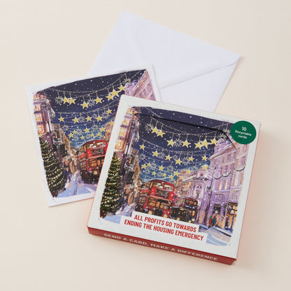 A boxed holiday card set with two illustrated cards and white envelopes on a light beige background. The front box shows a snowy city street scene with red buses, decorated trees, and starry night sky, plus text reading 'ALL PROFITS GO TOWARDS ENDING THE HOUSING EMERGENCY' and a green circular badge that says '10 illustrated cards.