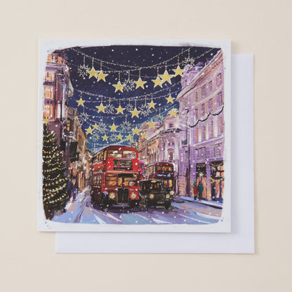 A square greeting card featuring a festive Christmas street scene with red double-decker buses, glowing yellow stars, and holiday lights, displayed on a white envelope against a light beige background.