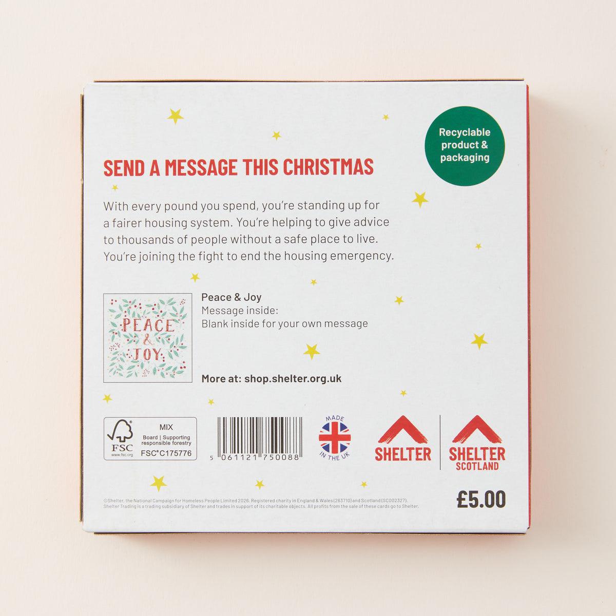 The back of a white rectangular box with red border and text, featuring holiday-themed messaging about housing support, a small illustrated card design, a barcode, and Shelter logos. The box is photographed against a warm off-white background.