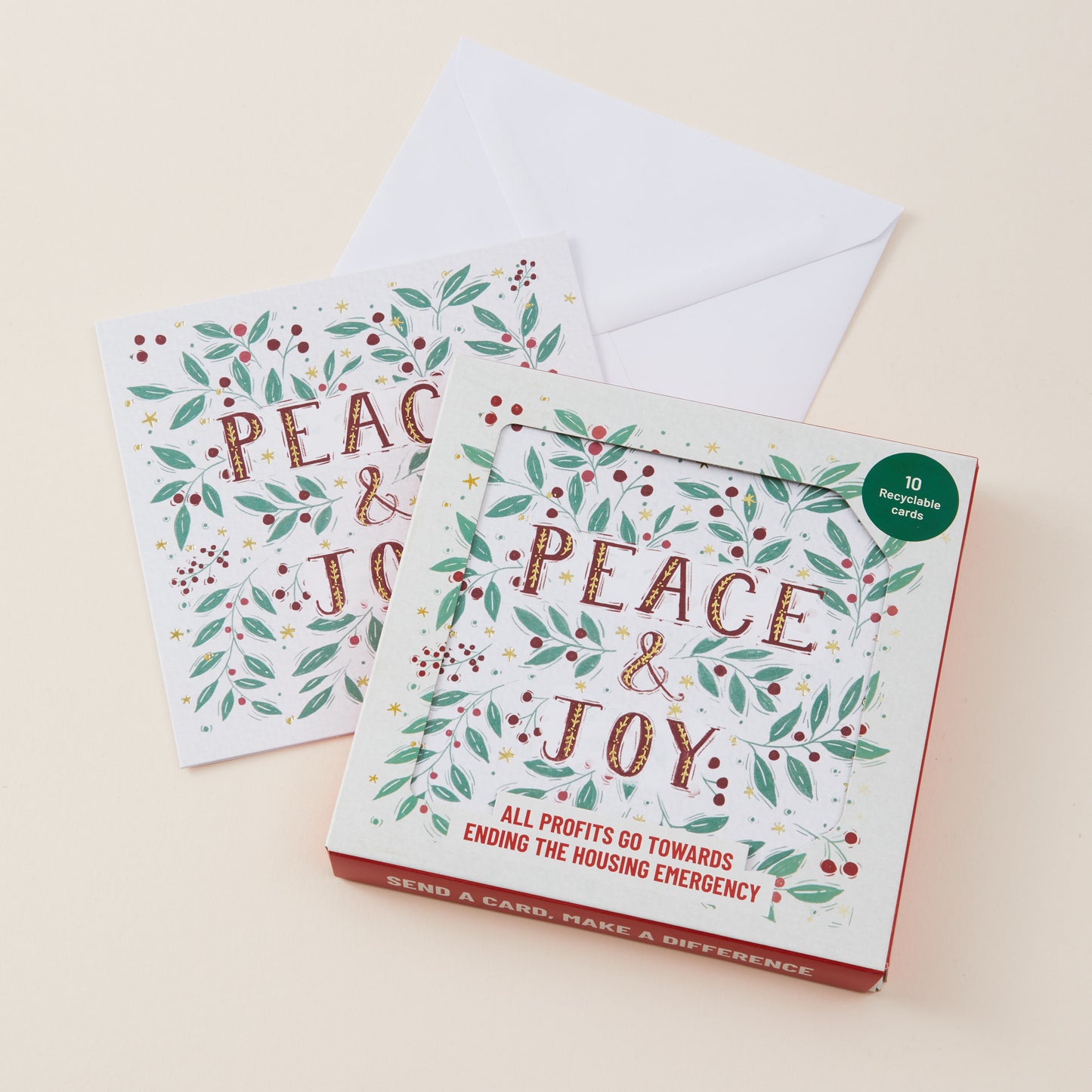 A boxed set of holiday greeting cards titled "PEACE & JOY" with two sample cards and an envelope on a warm cream background. The box has a white front, red border, green leaf and berry illustrations, and a small green circular badge reading "10 greeting cards.