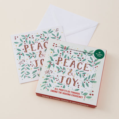 A boxed set of holiday greeting cards titled "PEACE & JOY" with two sample cards and an envelope on a warm cream background. The box has a white front, red border, green leaf and berry illustrations, and a small green circular badge reading "10 greeting cards.