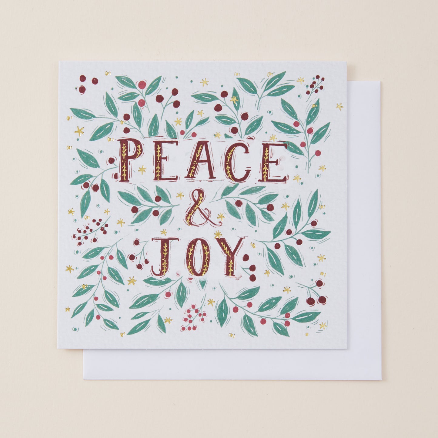 A square white greeting card with the words “PEACE & JOY” in textured brown lettering, surrounded by a festive wreath of green leaves, red berries, and small gold stars, shown with a white envelope on a light beige background.