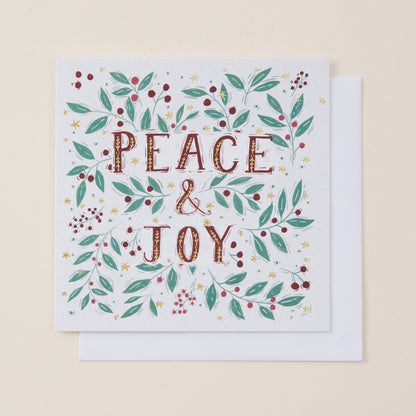 A square white greeting card with the words “PEACE & JOY” in textured brown lettering, surrounded by a festive wreath of green leaves, red berries, and small gold stars, shown with a white envelope on a light beige background.