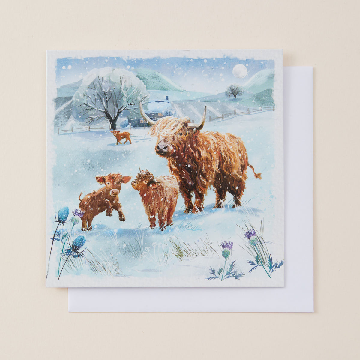 A square greeting card featuring a watercolor-style winter scene with Highland cows and calves in a snowy landscape, shown with a white envelope on a light beige background.